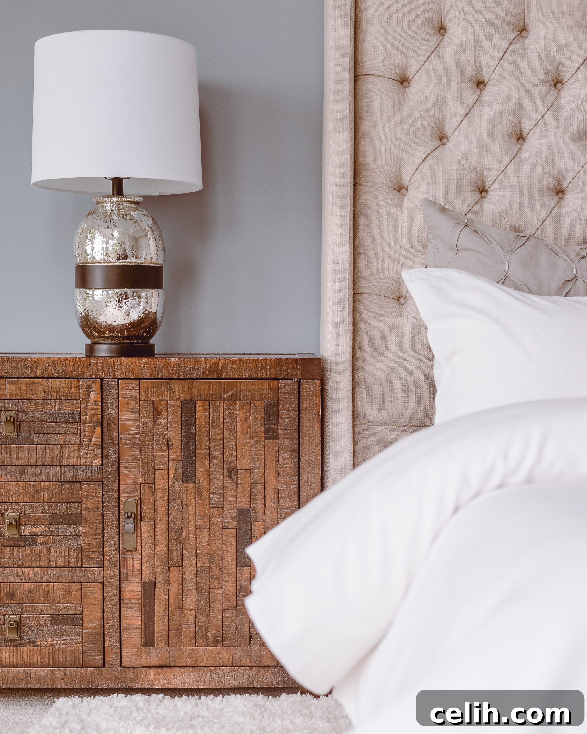 A bed with white linens and an upholstered headboard with a warm wood side table and metallic lamp